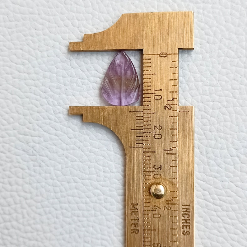May include: A purple amethyst gemstone carved into a leaf shape, measured with a ruler in inches. The gemstone is approximately 1 inch long.