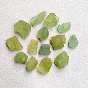 May include: A collection of 13 rough, green and yellow gemstones. The stones are various shapes and sizes, and have a natural, unpolished look.