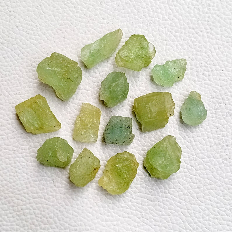 May include: A collection of 13 rough, green and yellow gemstones. The stones are various shapes and sizes, and have a natural, unpolished look.