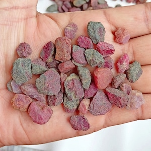 May include: A collection of rough, red ruby gemstones. The stones are various sizes and shapes, with some having a darker, almost black, hue.