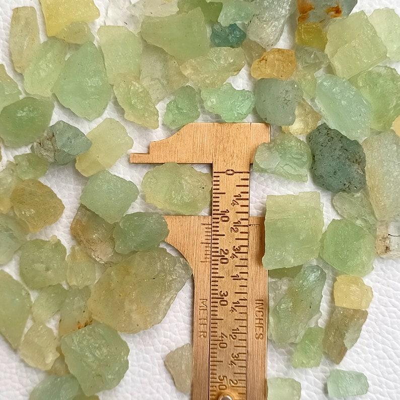May include: A close-up of a collection of green and yellow gemstone chips, measured with a ruler in inches. The chips are various sizes and shapes.