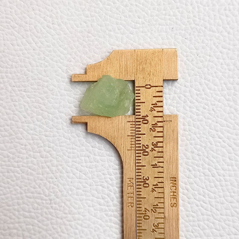 May include: A light green crystal with a rough, irregular shape, measured against a ruler in inches. The crystal is approximately 1/2 inch in size.