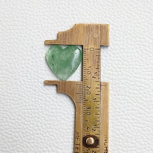 May include: A green heart-shaped stone being measured with a brass ruler. The stone is approximately 1 inch in length.