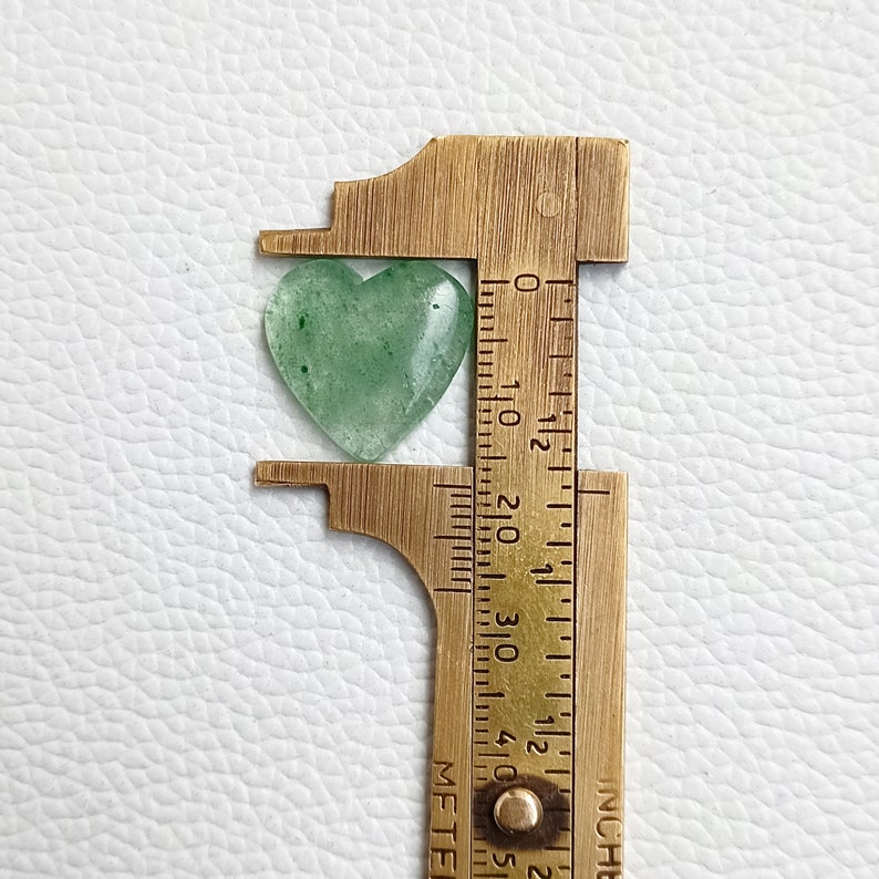 May include: A green heart-shaped stone being measured with a brass ruler. The stone is approximately 1 inch in length.