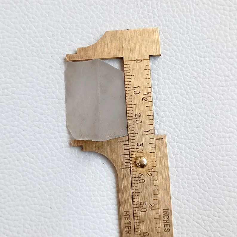 May include: A translucent, light gray gemstone is measured with a brass caliper. The caliper shows the stone is approximately 1.2 inches in length. The stone has a rough, irregular shape and is set against a white surface.