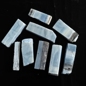 May include: A collection of rectangular, raw gemstone pieces. The stones have a pale blue hue with white and dark gray banding. The pieces are arranged on a black surface, showcasing their natural textures and colors. The gemstones appear to be cut from a larger stone.