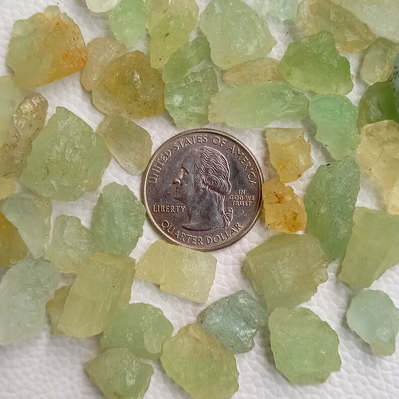 May include: A collection of small, green, and yellow, translucent, rough-cut gemstones, scattered on a white surface. A US quarter is included for scale.