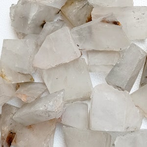 May include: A collection of raw, clear quartz crystal chunks. The crystals vary in size and shape, with a translucent appearance and subtle variations in color, ranging from clear to milky white. Some pieces have small, natural inclusions.