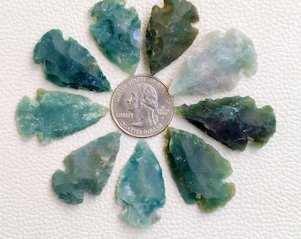 Puede incluir: Una colección de nueve piedras en forma de punta de flecha de color verde, marrón y blanco dispuestas en círculo alrededor de una moneda de un cuarto de dólar estadounidense. Las piedras son de varios tonos de verde y marrón con vetas blancas.
