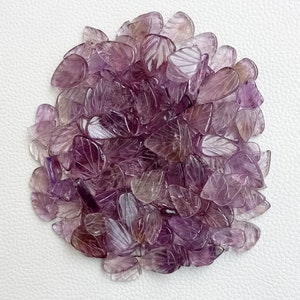 May include: A pile of purple amethyst leaf shaped gemstone beads. The beads are carved with a leaf design and have a translucent appearance.