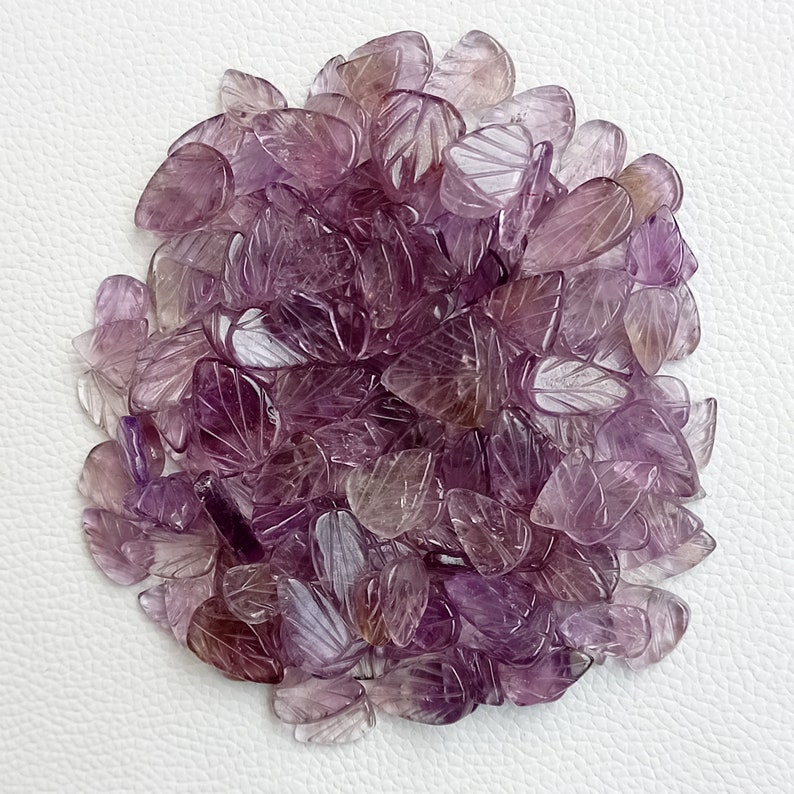 May include: A pile of purple amethyst leaf shaped gemstone beads. The beads are carved with a leaf design and have a translucent appearance.
