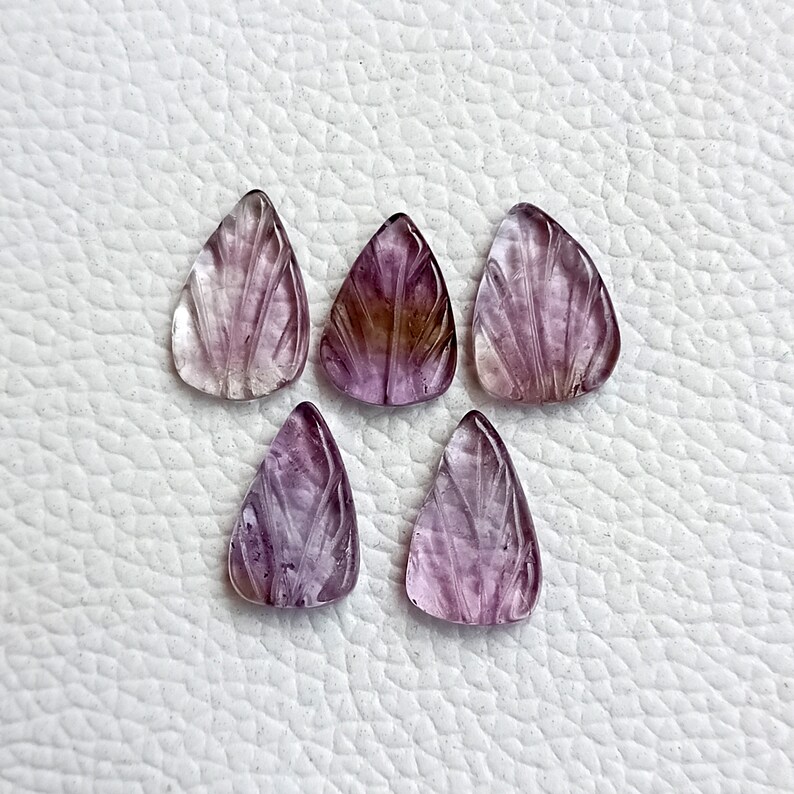 May include: Six purple and clear carved gemstone pendants in a teardrop shape. The pendants have a leaf design carved into them.