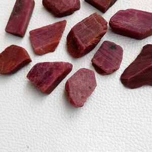 May include: A collection of ten rough, red ruby gemstones. The stones are various shapes and sizes, with some having a more rectangular shape and others being more irregular.
