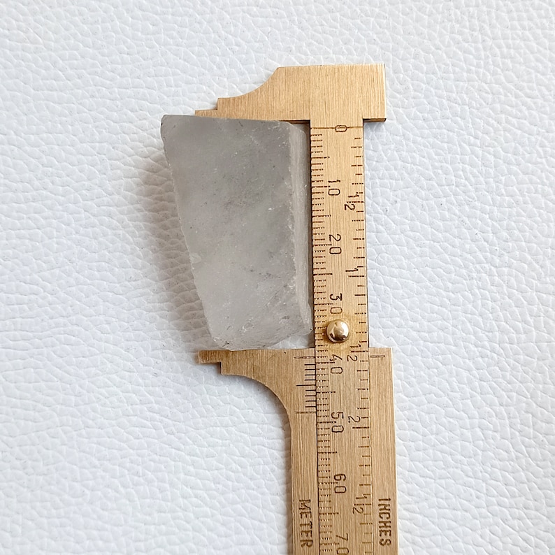 May include: A translucent, light gray rectangular stone is measured with a brass ruler. The ruler shows measurements in inches, with the stone appearing to be approximately 2 inches long. The stone has a rough, natural texture.