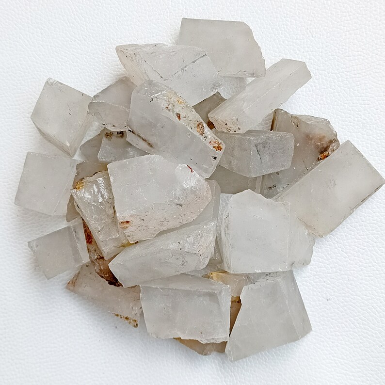 May include: A collection of raw, clear quartz crystal chunks. The crystals have a rough, natural texture and varying sizes. Some pieces have small, brown inclusions. The crystals are scattered on a white surface.