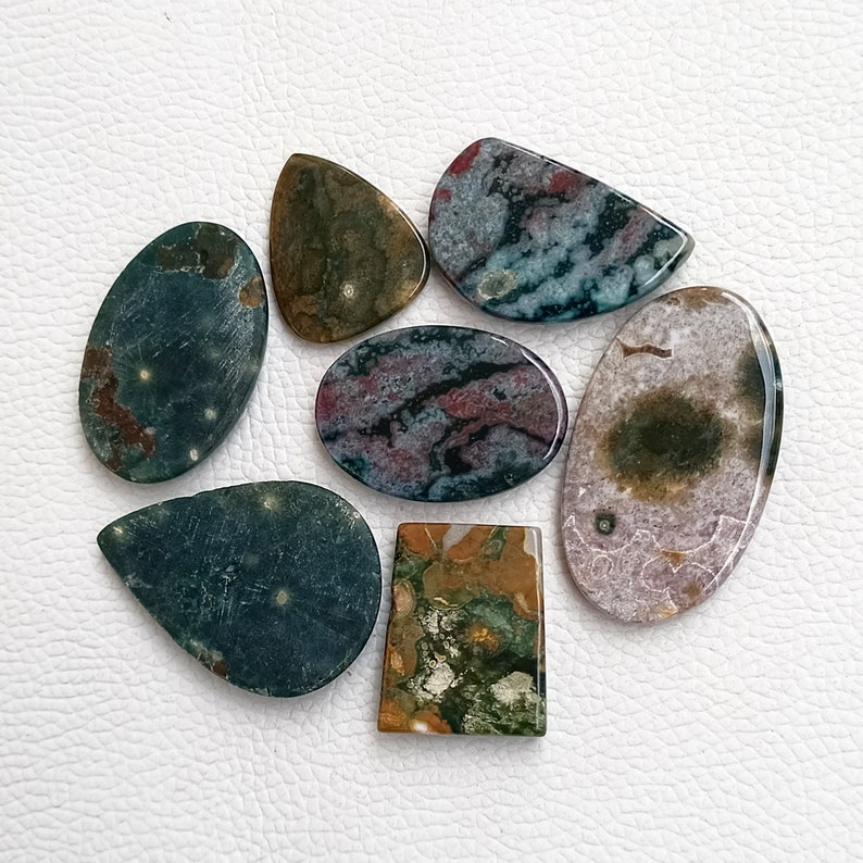 May include: Seven polished, irregularly shaped, green, brown, and pink stones with a marbled pattern. The stones are arranged on a white surface.
