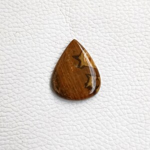 May include: A teardrop-shaped cabochon gemstone with a brown and white pattern. The stone has a smooth, polished surface.