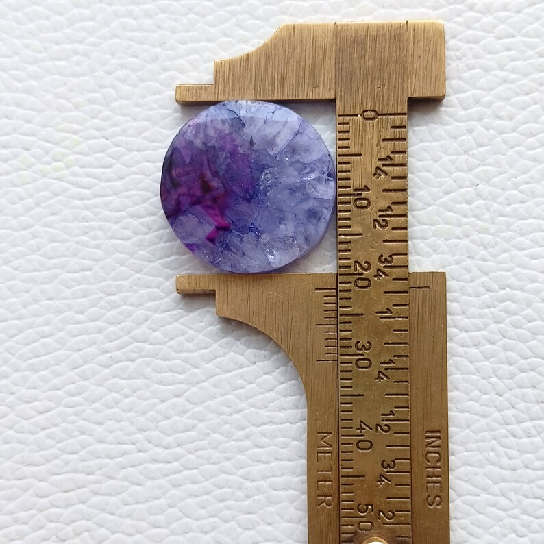 May include: A purple and white agate cabochon measuring approximately 1 inch in diameter. The cabochon is being measured with a ruler.