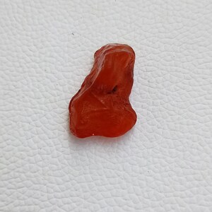 Op de afbeelding: Een kleine, onregelmatig gevormde, rode carneol edelsteen. De carneol is doorschijnend en heeft een glad, gepolijst oppervlak.