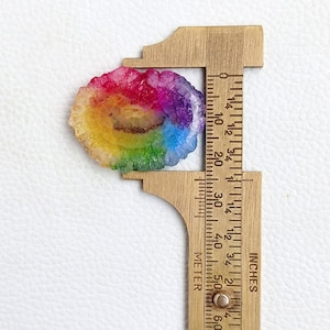 May include: A rainbow agate geode slice measuring approximately 1 1/4 inches in diameter. The geode has a natural, irregular shape and features a variety of colors, including red, orange, yellow, green, blue, and purple.