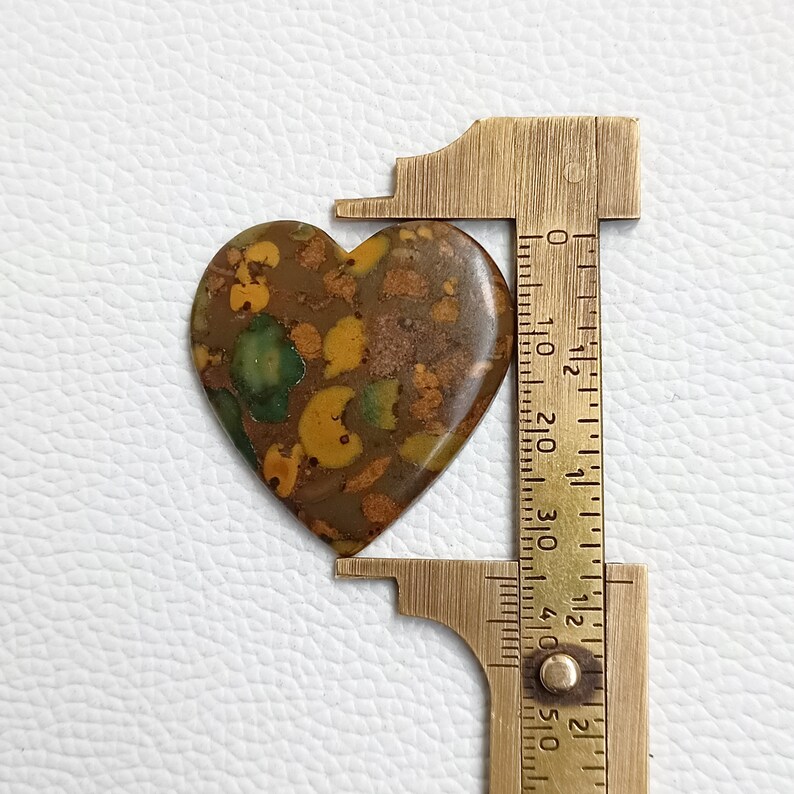 May include: A heart-shaped, brown, green, and yellow jasper cabochon, measuring approximately 2.5 cm in length, is being measured with a caliper.