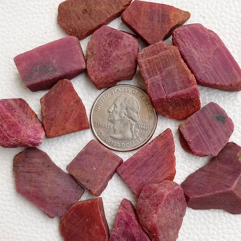 May include: A collection of 15 rough ruby gemstone pieces in various shapes and sizes. The gemstones are a deep red colour and have a natural, unpolished finish. A UK 20 pence coin is included for scale.