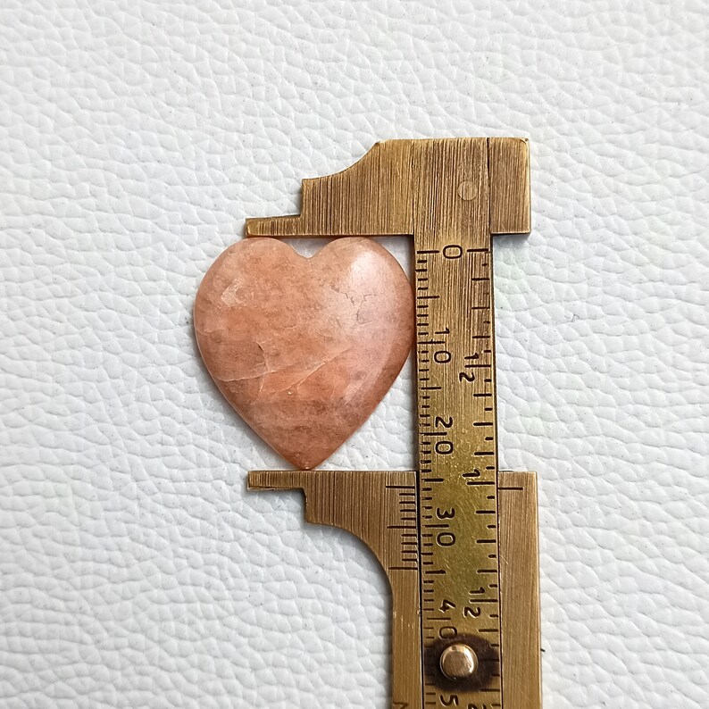 May include: A heart-shaped, peach-colored gemstone with white veins, measured with a ruler. The gemstone is approximately 2.5 cm wide.