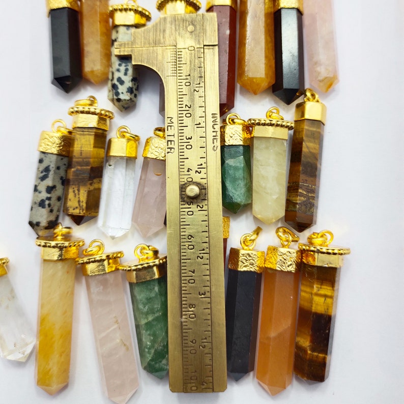 May include: A collection of 16 gold-capped crystal point pendants, each with a different colour and pattern. The pendants are arranged in a row and measured with a ruler. The ruler is marked in inches and centimetres.