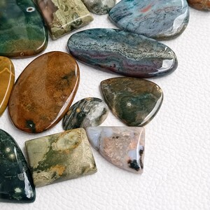 May include: Assortment of polished green, brown, blue, and gray ocean jasper stones. The stones are various shapes and sizes, including oval, teardrop, and rectangular.