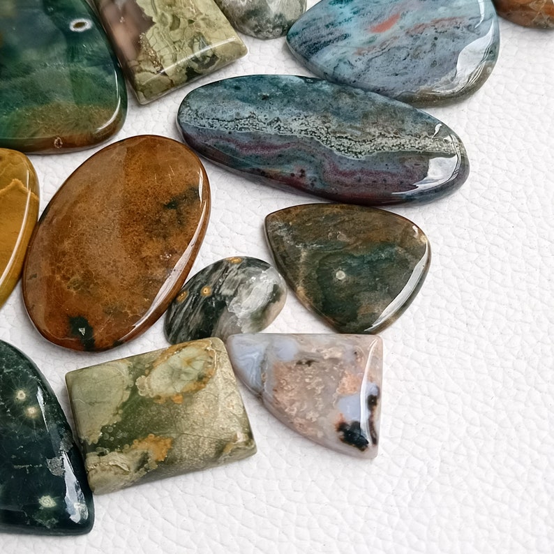 May include: Assortment of polished green, brown, blue, and gray ocean jasper stones. The stones are various shapes and sizes, including oval, teardrop, and rectangular.