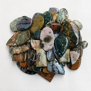 May include: A collection of polished green, brown, and white ocean jasper cabochons. The stones are various shapes and sizes, with some having a speckled pattern.
