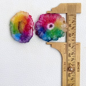 May include: Two rainbow agate slices, each with a different color pattern. The slices are approximately 1 inch in diameter and are measured with a ruler.