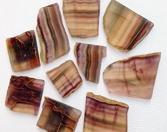 May include: Ten pieces of raw, multi-colored fluorite. The fluorite is banded with shades of purple, green, brown, and white. The fluorite is irregularly shaped and has a natural, rough texture.