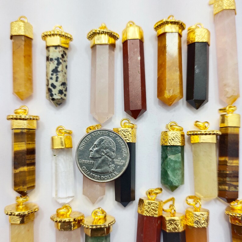 May include: Assortment of 17 gold-capped crystal point pendants in various colours and patterns, including tiger's eye, clear quartz, rose quartz, black onyx, green aventurine, and more. The pendants are arranged on a white surface with a US quarter dollar for scale.
