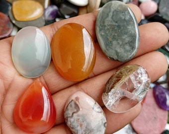 May include: Assortment of six polished oval and teardrop shaped gemstones in various colors, including green, orange, white, and red. The stones are displayed on a hand.