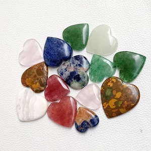 May include: Assortment of heart-shaped polished stones in various colors, including pink, green, blue, brown, and white. The stones are arranged on a white surface.