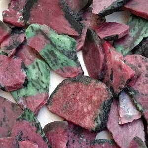 Może przedstawiać: Kolekcja surowych, naturalnych zielonych i r&oacute;żowych kamieni szlachetnych. Kamienie mają r&oacute;żne kształty i rozmiary, a niekt&oacute;re mają czarne oznaczenia.