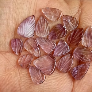 May include: A collection of 18 purple amethyst gemstone beads, each carved with a leaf design. The beads are smooth and polished, with a translucent appearance.