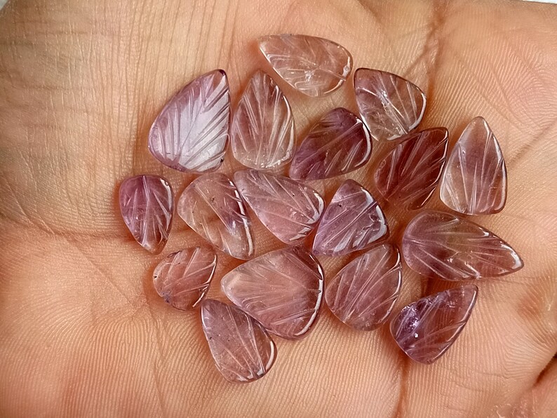 May include: A collection of 18 purple amethyst gemstone beads, each carved with a leaf design. The beads are smooth and polished, with a translucent appearance.