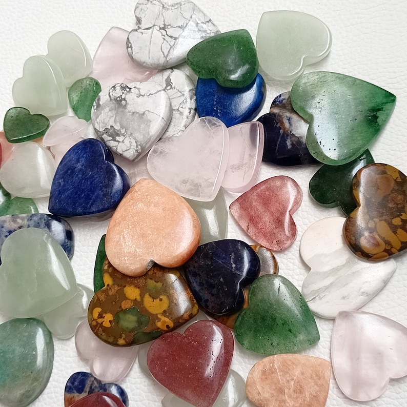 May include: A collection of heart-shaped polished stones in various colors, including green, blue, pink, brown, and white. The stones are arranged in a random pattern on a white surface.