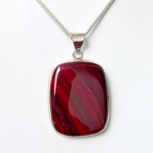 May include: A silver chain necklace with a red agate pendant. The pendant is a rectangular shape with a smooth, polished surface. The agate has a swirling pattern of red and white.