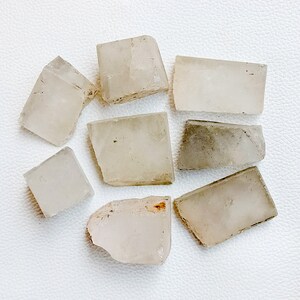 May include: A collection of eight raw, translucent calcite crystal chunks. The crystals vary in size and shape, with a pale, off-white color and rough, fractured edges. Some pieces have slight variations in tone, with hints of gray or tan.