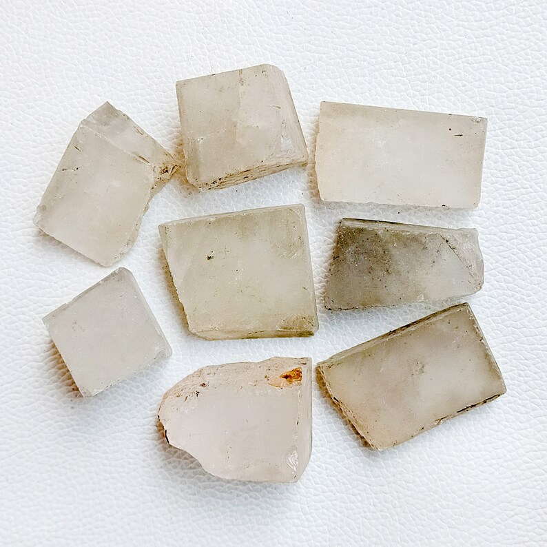 May include: A collection of eight raw, translucent calcite crystal chunks. The crystals vary in size and shape, with a pale, off-white color and rough, fractured edges. Some pieces have slight variations in tone, with hints of gray or tan.