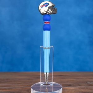 May include: A light blue ballpoint pen featuring a Buffalo Bills football helmet topper, two round blue beads, and a single red rhinestone spacer bead. The pen is displayed upright in a clear acrylic stand on a wooden surface.