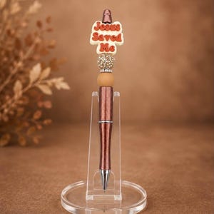 May include: A metallic brown ballpoint pen featuring a decorative topper with the text Jesus Saved Me in red letters on a white background, accented by a gold glitter bead, a silver rhinestone spacer, and a smooth tan bead on a clear stand.