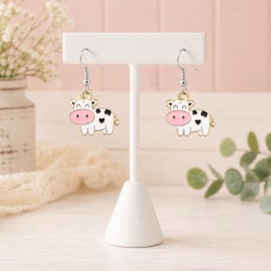 May include: A pair of dangle earrings featuring white cow charms with pink snouts and black spots. The charms are suspended from silver-toned metal hooks. Each cow charm has a small black heart detail on its side, creating a cohesive look.
