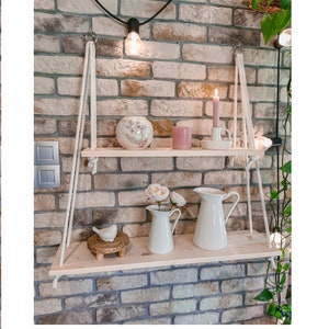Op de afbeelding: Twee houten planken die aan een bakstenen muur hangen met touw. De planken zijn versierd met witte keramische kannen, een roze keramische vaas met roze bloemen, een wit keramiek vogeltje en kaarsen.