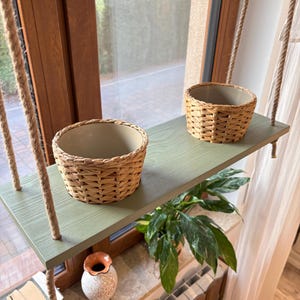 Könnte beinhalten: Ein Hängeregal mit zwei geflochtenen Körben, einem grün lackierten Holzregal und einem Seil, das von der Decke hängt. Das Regal ist in der Nähe eines Fensters mit einer Pflanze auf der Fensterbank montiert.