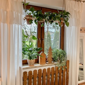 Pode incluir: Uma janela com cortinas brancas e uma cobertura de radiador de madeira. Há plantas penduradas em vasos de terracota e uma pequena casa de madeira e uma decoração de árvore de Natal na soleira da janela.