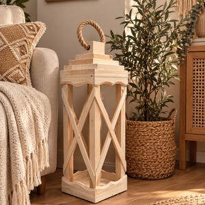Può includere: Una lanterna quadrata in legno alto con una finitura in legno naturale chiaro. La struttura presenta una parte superiore a gradini, una spessa maniglia in corda di juta e pannelli laterali decorati con un motivo a croce. La base e i bordi hanno finiture smerlate.